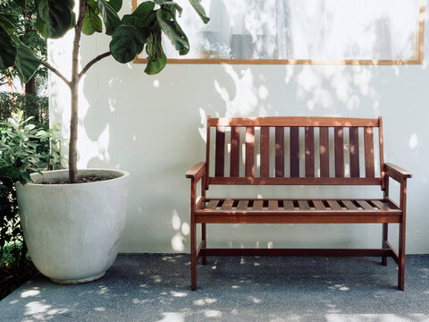 Empty Minimal Style Cozy Wooden Bench With Arms Decorated On A White Wall Near The Big Concrete Pot Outside The Building Near A Window And The Green Garden.