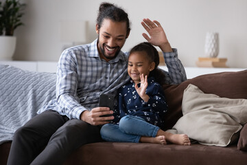 Happy Afro American millennial dad and cheerful cute kid talking on video call together, waving hand hello at smartphone screen, using telephone for online conference. Family communication concept