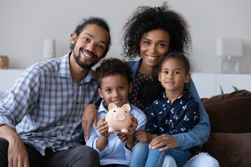 Happy Black family couple and little kids making financial reserve, donation funk, keeping money in piggybank, showing moneybox, looking at camera, smiling. Head shot home family portrait