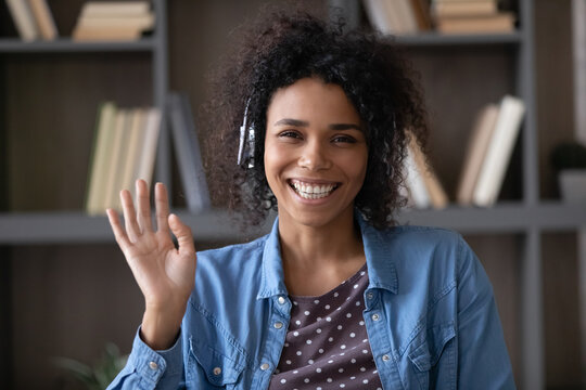 Happy excited African student, employee in wireless headphones waving hand hello with toothy smile, laughing at camera, making video call at home. Black woman talking online head shot portrait screen