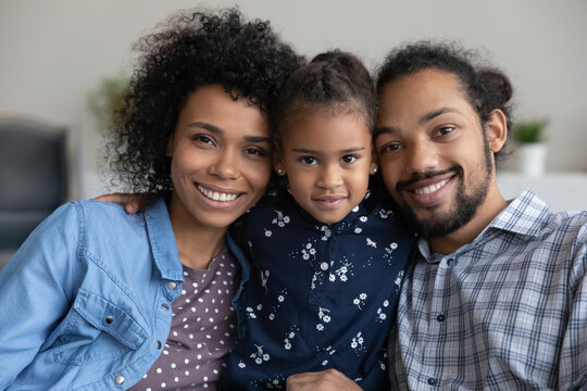 Happy Black millennial family couple holding adorable little daughter child in arms, looking at camera with toothy smiles. Pretty preschool girl hugging parents. Home head shot portrait