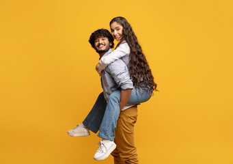 Young indian man giving piggyback ride to his girlfriend against yellow studio background, free space