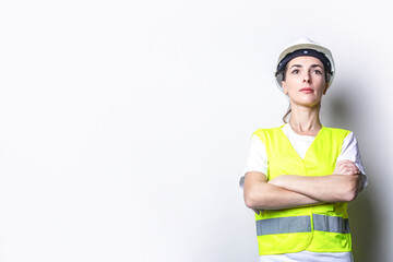 Young woman in building clothes in a hard hat with folded hands on a light background.