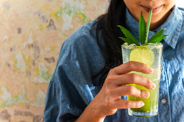 An Asian woman in blue jeans shirt sip or drinks cannabis lemon soda over abstract colorful wall in...
