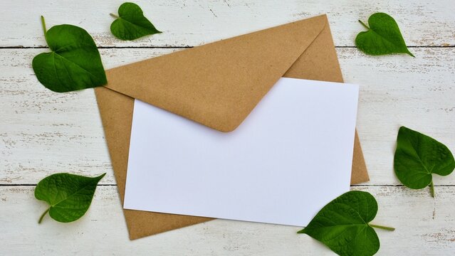 Mockup of a kraft paper envelope with  white card with green leaves in the shape of a heart on a white wooden background  closeup. Valentines Day, wedding, Mother's Day greeting card 