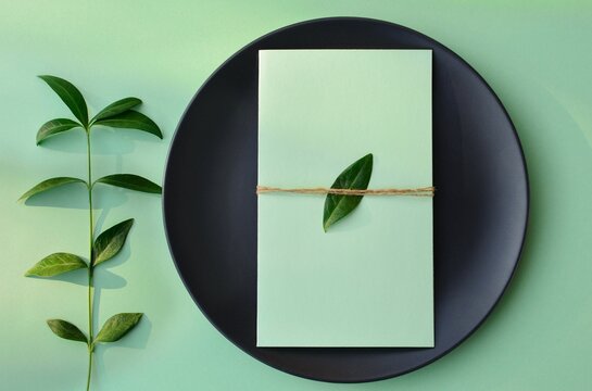  Mockup of black plate with light green invitation card on green background with fresh leaves of a plant closeup.