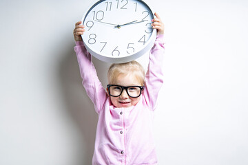 Beautiful child girl in glasses holds a large wall clock on a light background.
