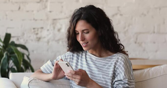 Cheery Latina woman celebrate online victory sit on sofa at home, holds cellphone check e-mail app on device feel excited looking happy by lottery win. Winner enjoy moment of success, betting concept