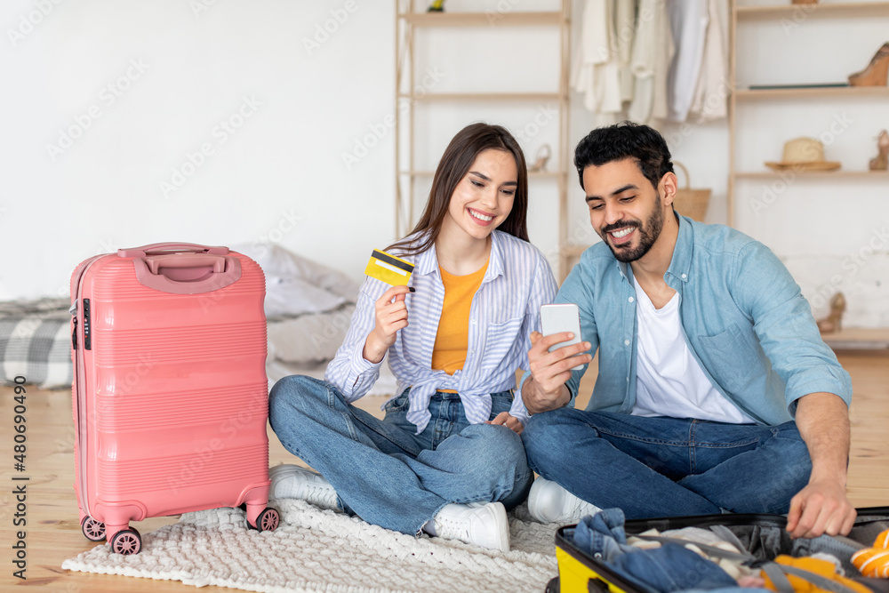 Wall mural Happy travelers couple using smartphone and credit card for booking tickets online in travel app, sitting near suitcases