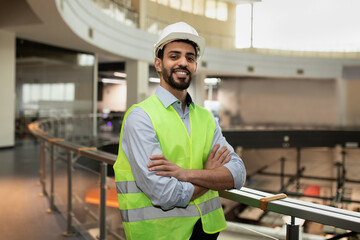 Happy confident young attractive arabic engineer in hard hat and protective clothing in office plant interior