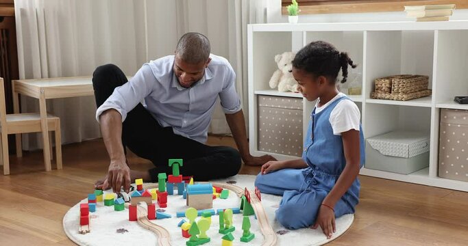 Happy Afro American father having fun with preteen girl move toy train by railroad built from bricks at nursery room. Single dad sit on warm carpet enjoy playing constructor set with tween daughter