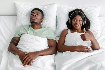 Young african american guy sleeping on bed, cheerful lady in headphones shows thumb up in bedroom,...