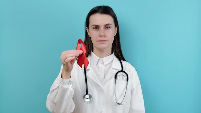 Doctor woman holds red ribbon, looking at camera, wears uniform and stethoscope, isolated on blue background studio. Blood transfusion and donation. Hemophilia, health concept. World AIDS and HIV day