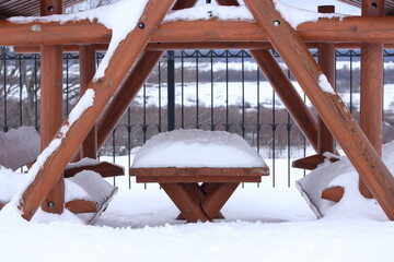 Snow on the table and benches of the gazebo against the background of a forged fence and meadows