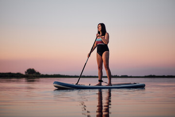 Relaxed middle aged woman rowing on sup board with oar looking at sunset on lake rippled water with pink sky in background. Active lifestyle for older people.