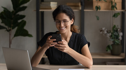 Happy millennial generation female employee in eyewear using telephone, distracted from laptop work in modern office. Smiling 30s young hispanic latina business lady using mobile applications.