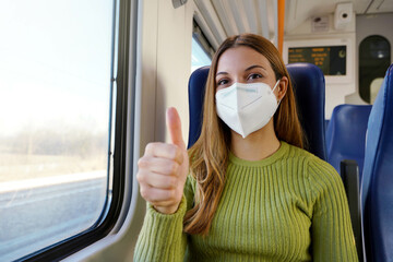 Optimistic young woman wearing protective medical face mask on public transport showing thumbs up...