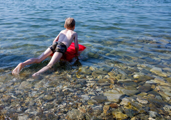 a boy is swimming in the sea on a red inflatable mattress.