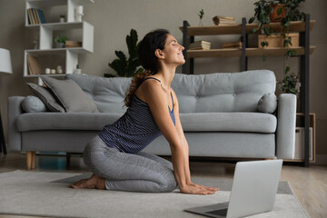 Smiling sporty young hispanic latin woman in active wear sitting on carpet floor, starting online...