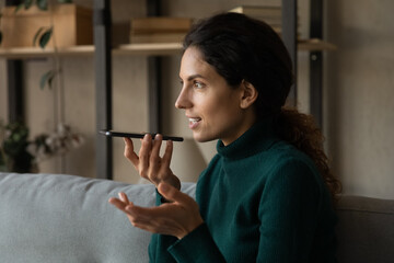 Joyful young hispanic woman holding smartphone in hands, dictating audio message, sending...