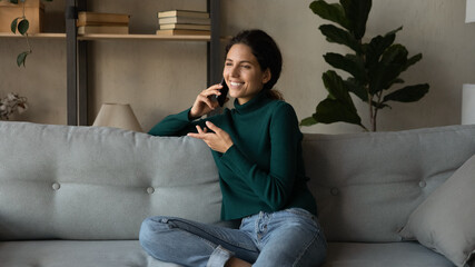 Fototapeta premium Smiling beautiful young woman involved in mobile phone call conversation with friends, talking chatting speaking communicating distantly resting on comfortable sofa alone in modern living room.