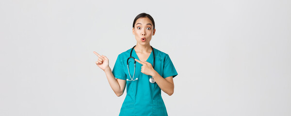 Covid-19, healthcare workers, pandemic concept. Impressed and amazed female asian nurse in scrubs asking question with intrigued expression. Doctor looking thrilled and pointing upper left corner - obrazy, fototapety, plakaty