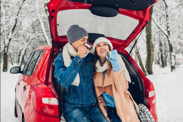Fototapeta Valentine's day. Couple in love sitting in car trunk drinking hot tea in snowy winter park. People relaxing outdoors obraz