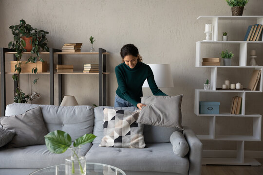 Happy millennial generation positive beautiful young housewife cleaning apartment, arranging stuff in cozy modern living room, put things on right places, feeling satisfied with comfort tidy house.