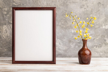 Wooden frame with yellow barrenwort flowers in ceramic vase on gray concrete background. side view, copy space, mockup.