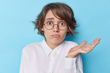 Puzzled hesitant young brunette European woman raises palm with clueless expression cannot make decision wears round spectacles and white formal shirt isolated over blue background. So what to do