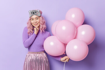 Obraz na płótnie Canvas Pleased pink haired Asian woman wears sleepmask jumper and pleated skirt holds inflated balloons celebrates birthday isolated over purple background. People special events and celebration concept