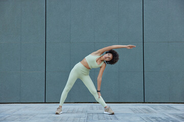 Fototapeta Horizontal shot of sporty slim woman in activewear leans aside exercises against blank grey wall outdoors stays healthy keeps good pysical form has regular training follow daily morning regime obraz