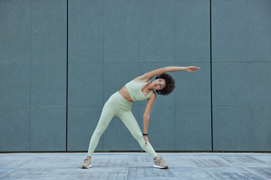 Horizontal shot of sporty slim woman in activewear leans aside exercises against blank grey wall outdoors stays healthy keeps good pysical form has regular training follow daily morning regime