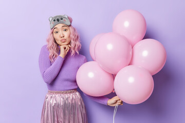 Obraz na płótnie Canvas Attractive Asian woman keeps lips rounded has tender face expression holds bunch of inflated balloons dressed in festive clothes comes on party isolated over purple background. Holiday concept