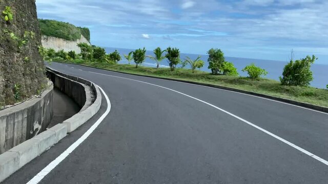 POV driving along the road with cavate rock on one side and blue ocean on the other. Seaside asphalt highway in tropical paradise. First person view riding along sea comb under blue sky.
