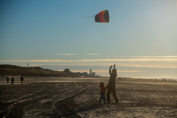 Father and son flying kite together on the beach in winter sunshine - obrazy, fototapety, plakaty