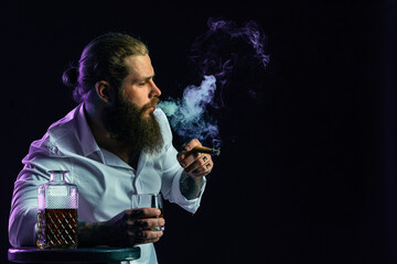 Handsome bearded sitting man smoke cigar holds glass of whiskey, dressed in white shirt. Studio shot