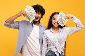 Rich millennial indian lovers hiding faces behind cash, holding bunches of dollars by faces over...
