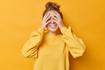 Carefree glad woman covers eyes with hands waits for surprise stads cheerful has hair combed in bun wears casual jumper isolated over yellow background plays hide and seek anticipates something good