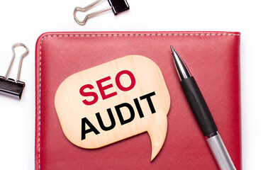 On a light background there are black paper clips, a pen, a burgundy notepad a wooden board with the text SEO AUDIT