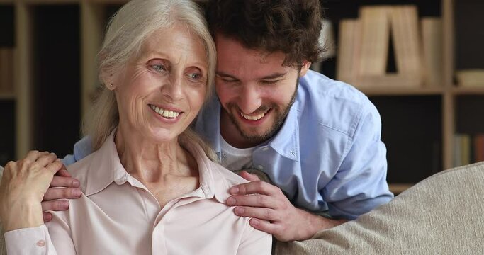 Close up portrait older 60s woman her young adult son. Grown up grandson spend time cuddle his mature grey haired granny sit on couch enjoy talk smile feel happy. Multi generation family ties concept