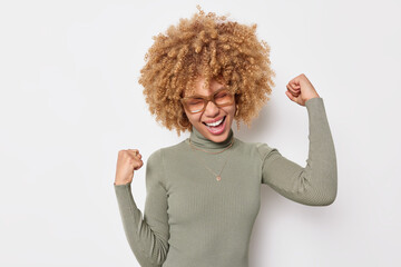 Overjoyed woman does winner gesture raises fists up celebrates achievements excaims happiy wears casual turtleneck transparent spectacles isolated over white background. People and success concept