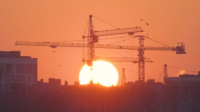 Dark silhouette of tower cranes with big setting sun at high residential apartment buildings construction site at sunset. Real estate development