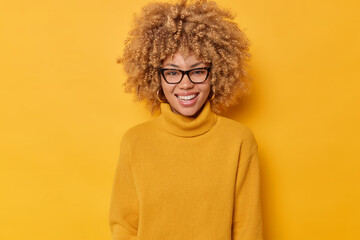 Fototapeta na wymiar Portrait of lovely young curly haired woman smiles happily shows white teeth wears spectacles and sweater looks directly at camera isolated over yellow background. Positive emotions concept.