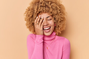 Positive curly woman covers face with palm giggles positively hears something funny shows white teeth wears pink turtleneck isolated over brown background. Happy emotions and feelings concept - obrazy, fototapety, plakaty