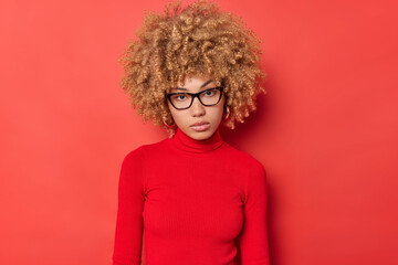 Portrait of serious female student with curly hair feels self confident gazes directly at camera...