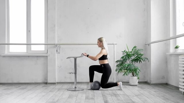Woman doing squats with fitness ball. Young sporty blonde training buttocks using ball in studio. Training and making physical exercises for legs and body indoors. 4K, UHD