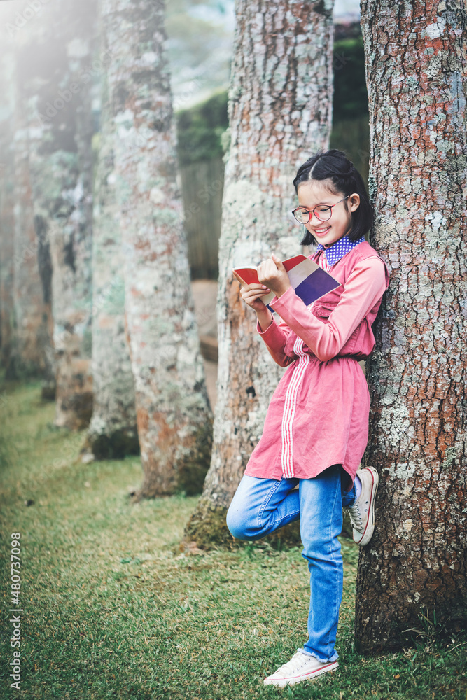 Wall mural Smiling asian cute little girl with glasses reading book in park