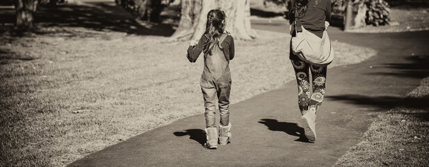 Mother and daughter walking along a beautiful city park on a sunny day, back view.