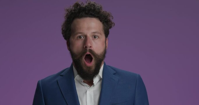 shocked bearded man in suit with curly hair opening mouth, being surprised, holding breath and making idiot faces, shaking head and being crazy on purple background